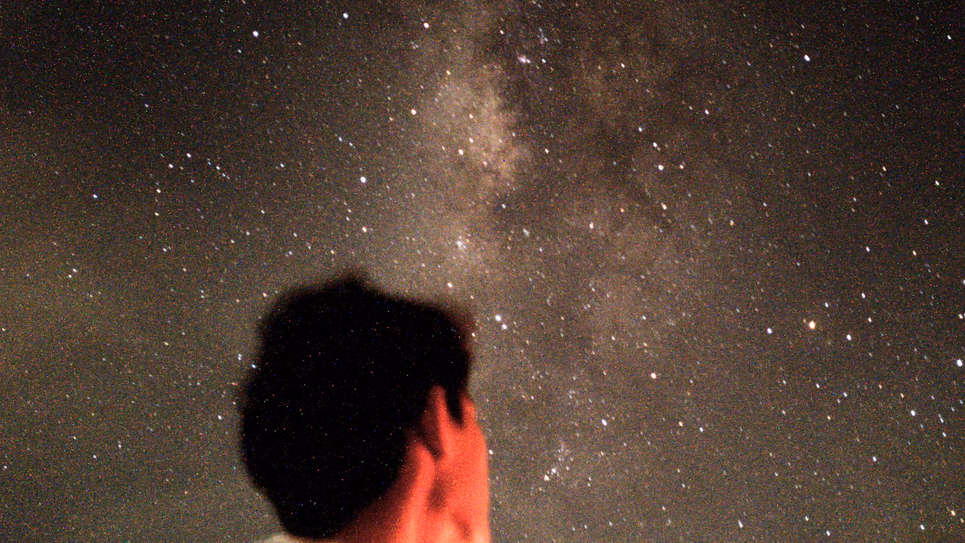A grainy, high-contrast shot of a person looking up at a brilliant Milky Way galaxy.