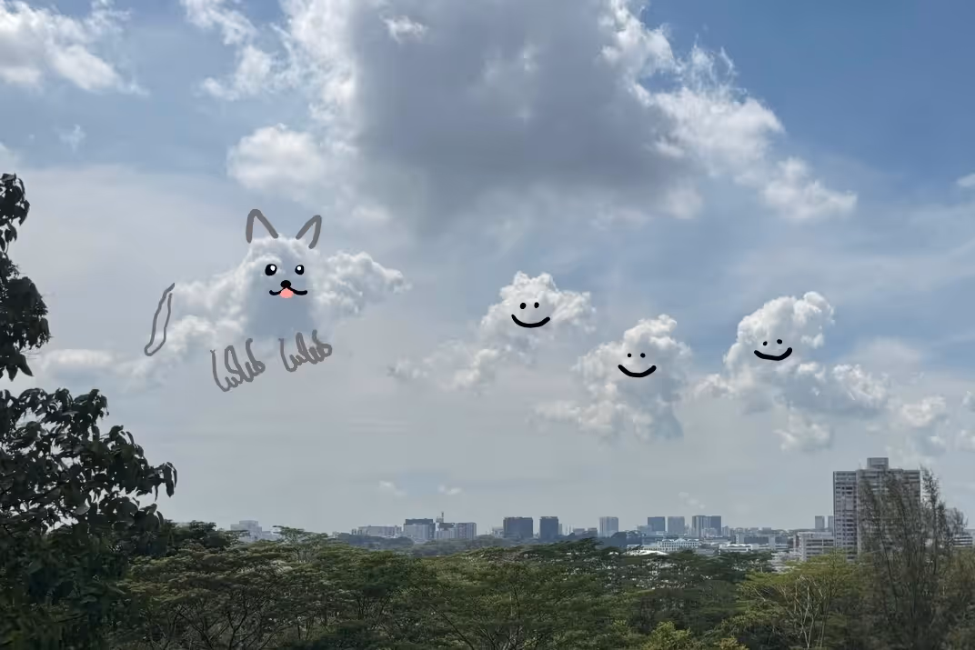 A wide landscape shot of a bright sky where a large cloud is edited to look like a cat with ears, paws, and a pink tongue, followed by three smaller clouds edited with simple black smiley faces, floating above a distant city skyline.