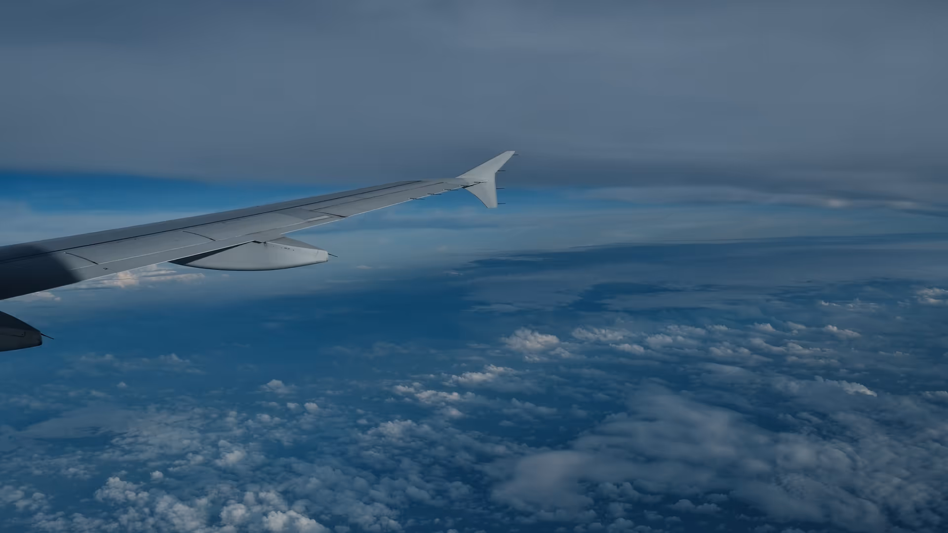 An airplane wing cutting through a sea of clouds at cruising altitude, blue sky above and a dense white cloud layer below.