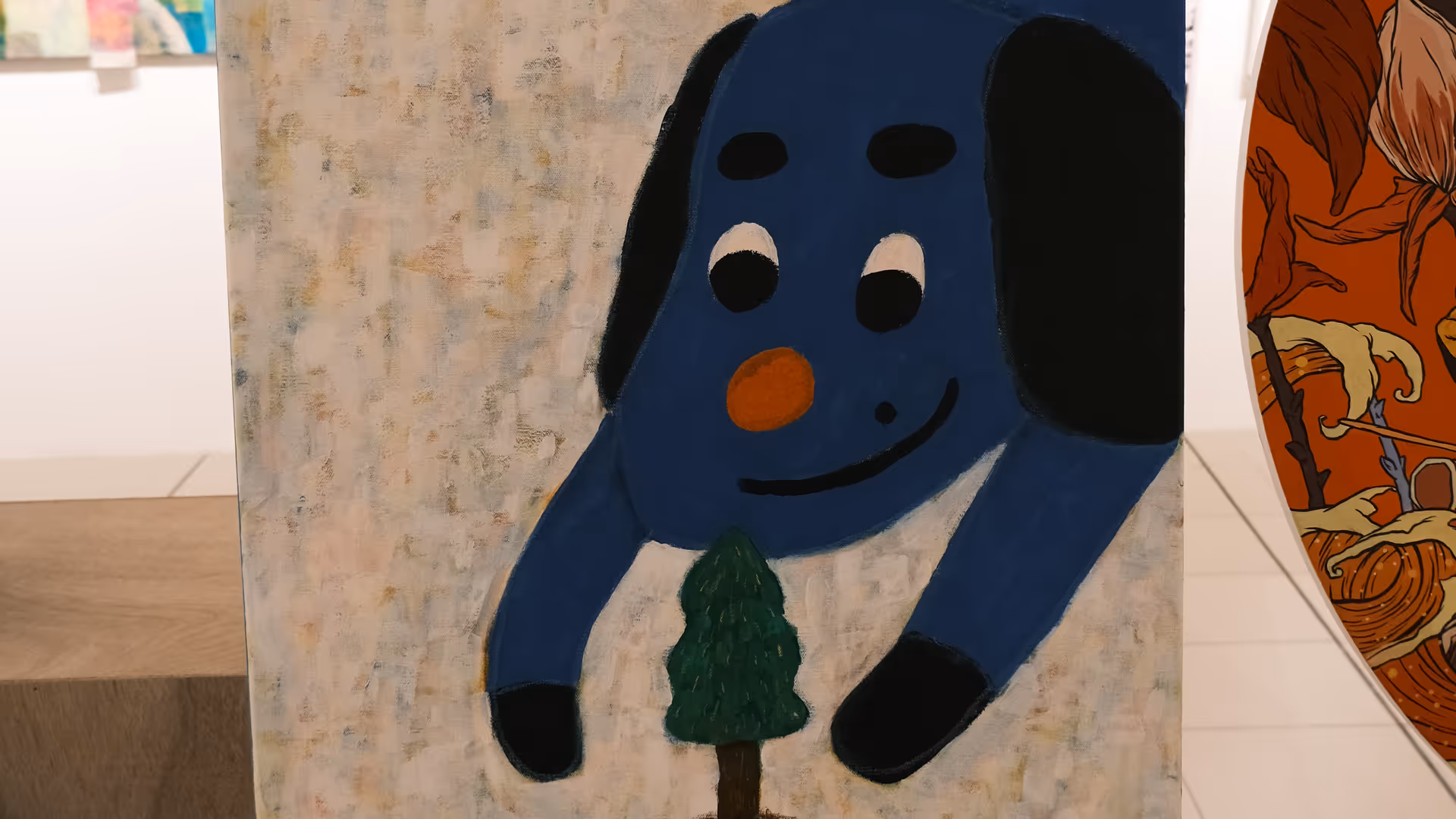 A large canvas painting of a cartoonish blue figure holding a small green plant, leaning on an easel surrounded by other artworks.