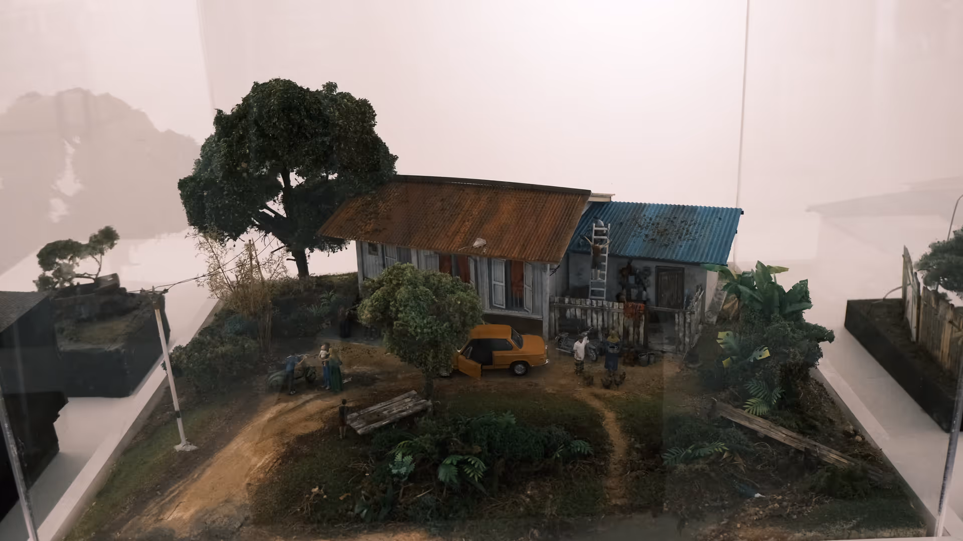 A detailed miniature kampung scene in a glass display case, complete with a zinc-roofed house, a yellow car, and lush tropical trees.
