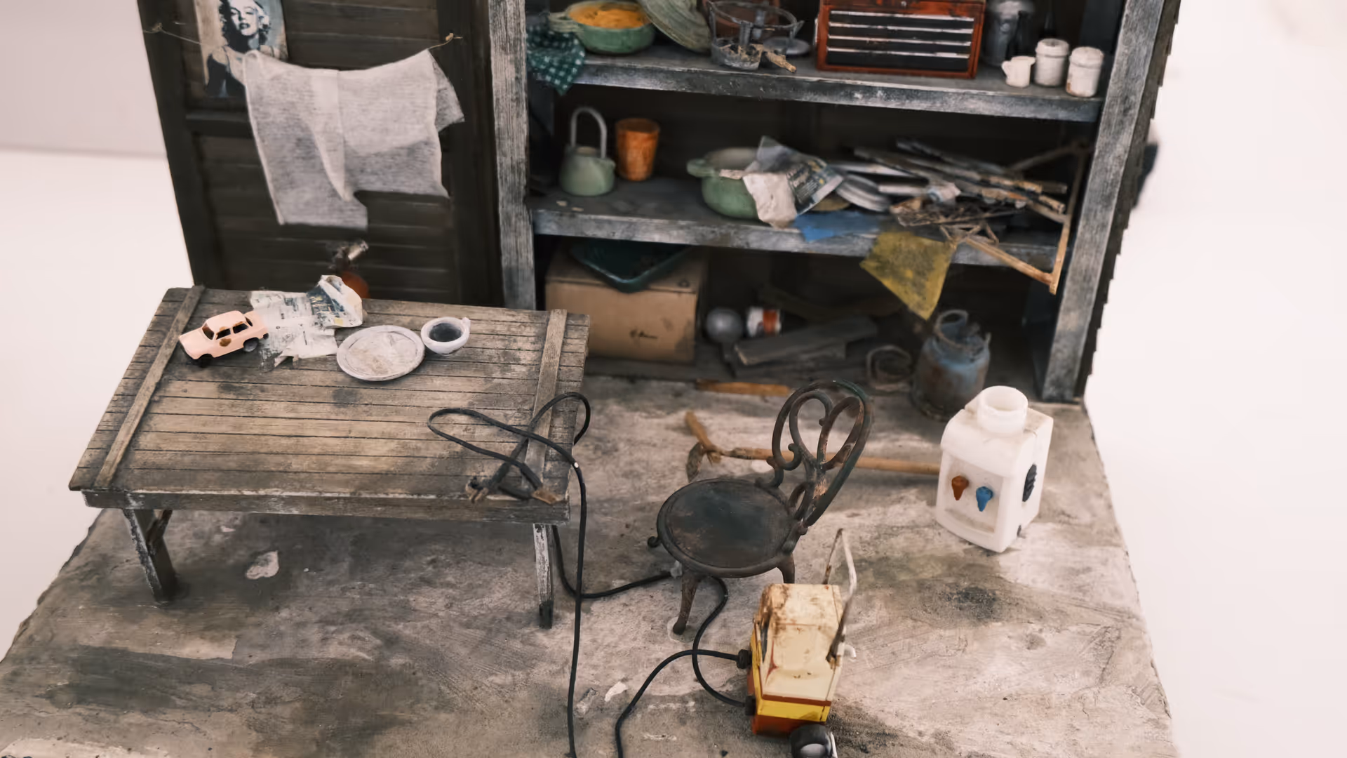A miniature diorama of a cluttered workshop with a wooden table, small iron chair, shelves full of tools, and a tiny toy truck on the floor.