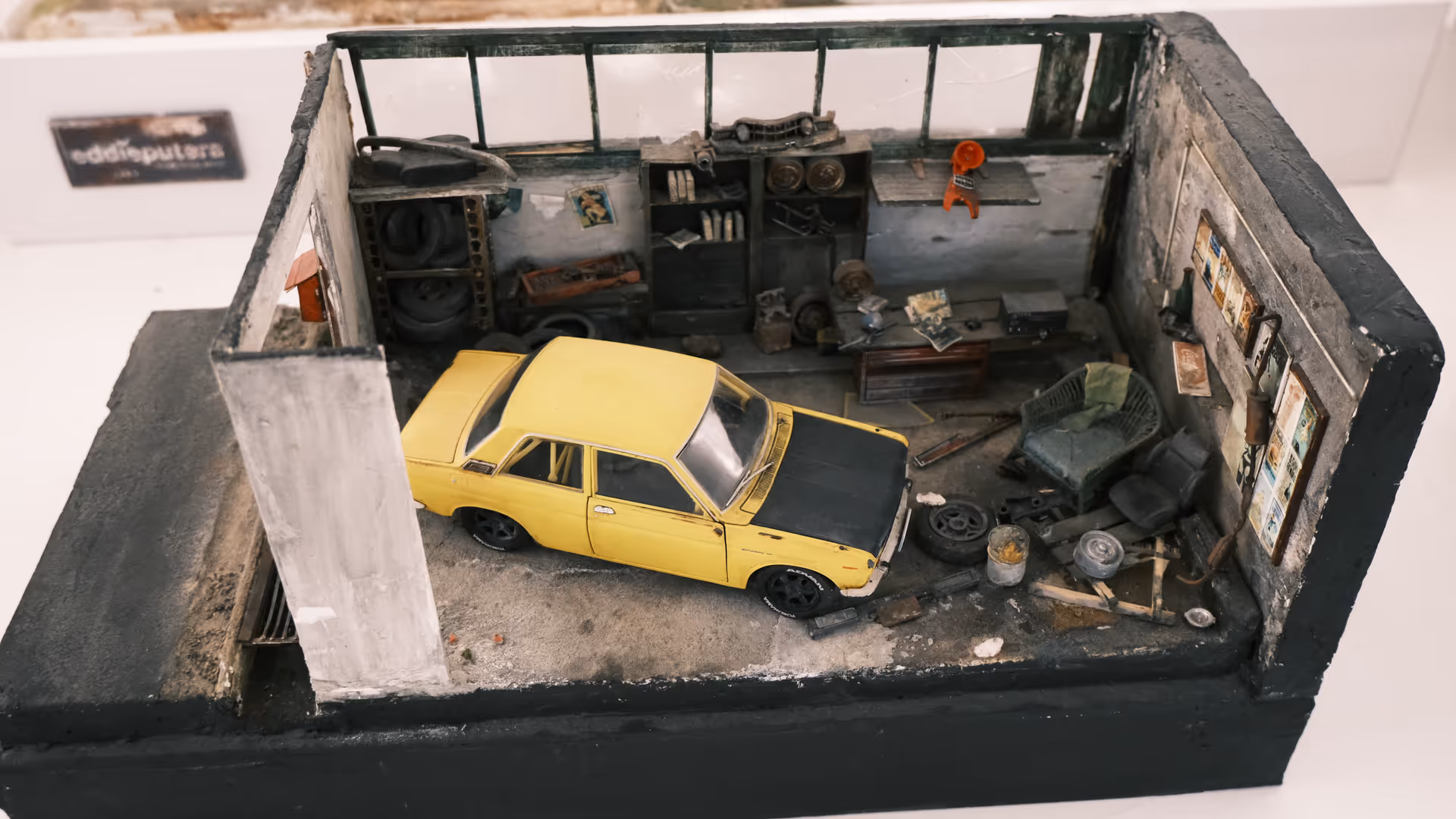 A highly detailed miniature diorama of a rundown mechanic's garage with a yellow classic car inside.