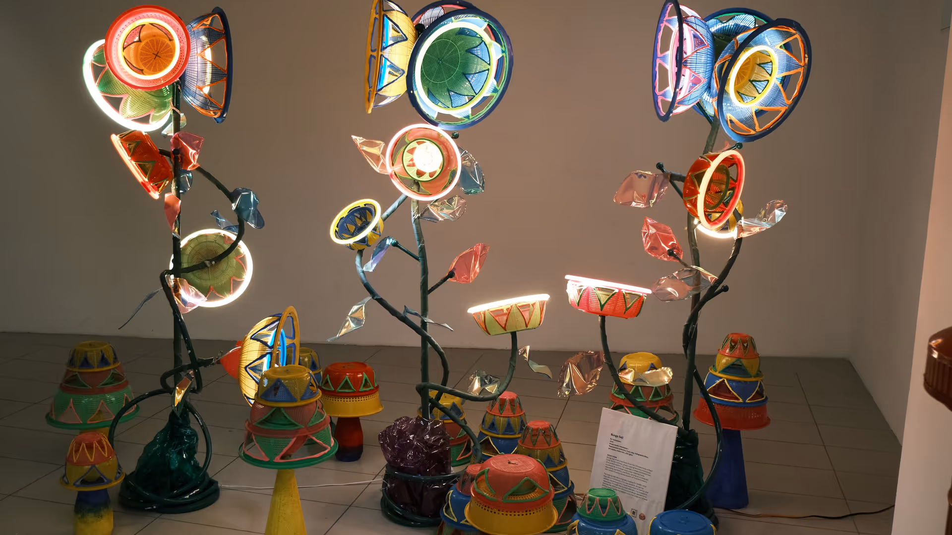 Colourful neon light sculptures shaped like flowers made from recycled pots and plates, glowing in a dim gallery space at GMBB.