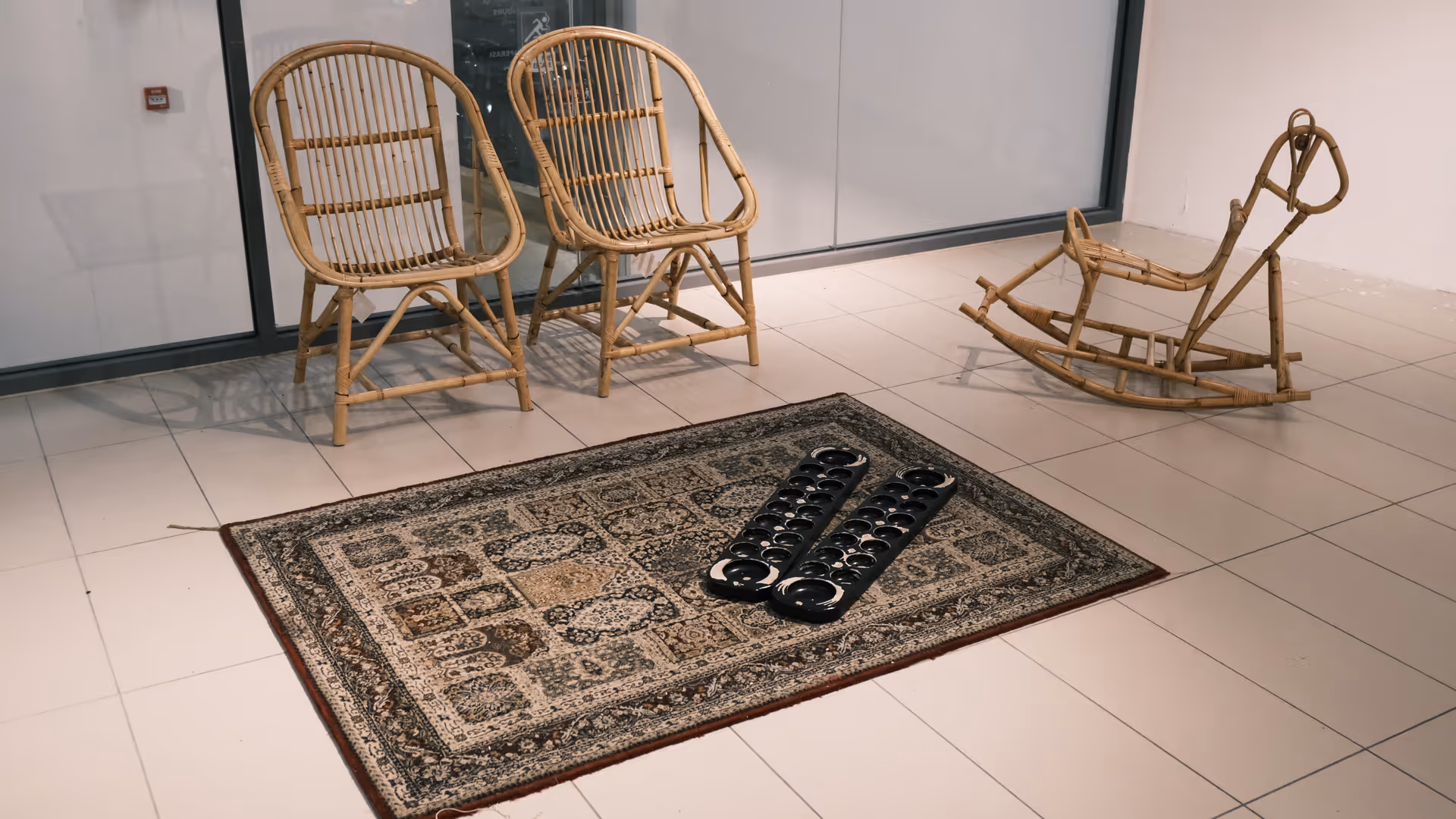 Rattan chairs and a rocking chair arranged around a patterned rug on a white gallery floor, with black objects laid on the carpet.