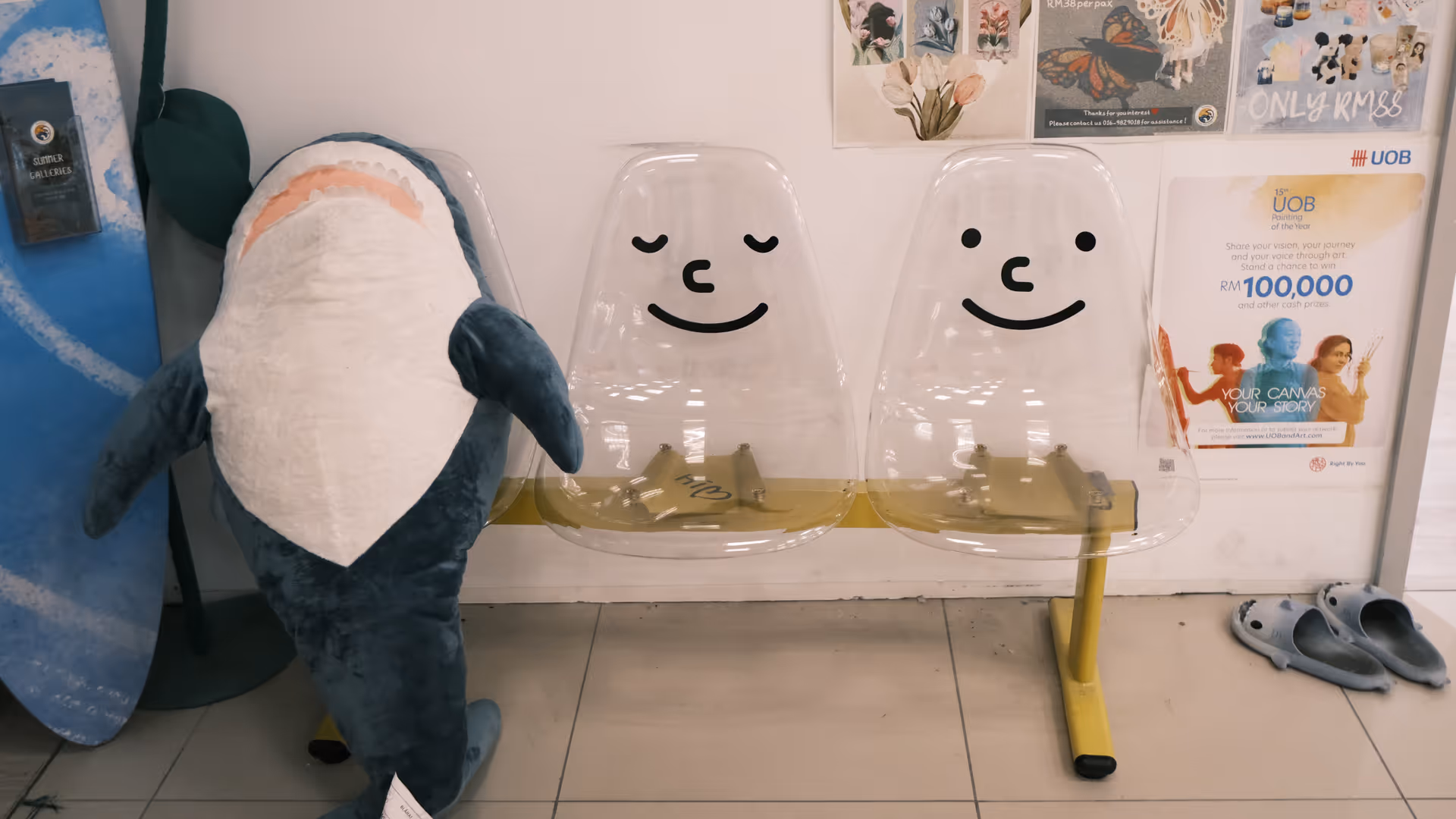 An IKEA shark plushie sitting on a bench with smiley face cutouts, next to a surfboard and gallery posters at GMBB.