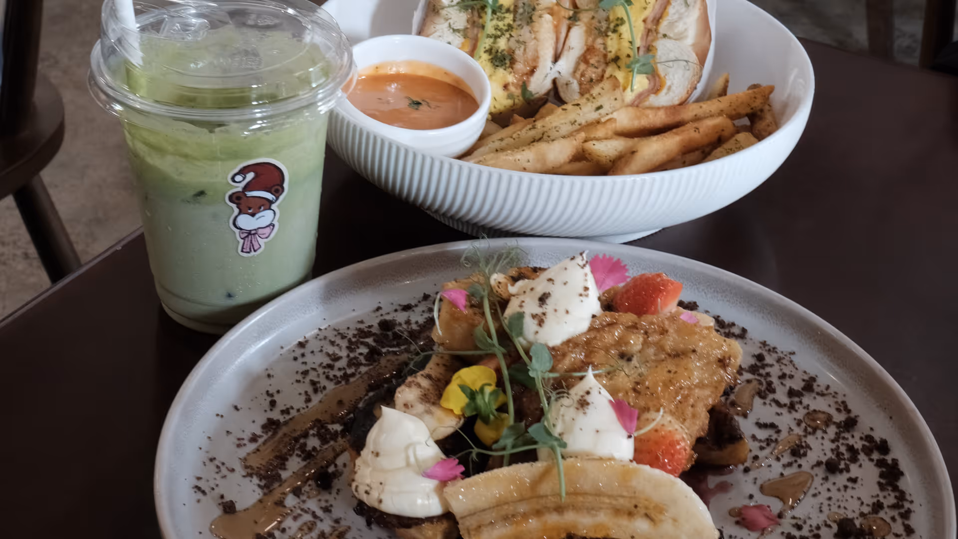 A spread of two bagel plates and a banana dessert with an iced matcha on a dark café table at Grumpy Bagels.