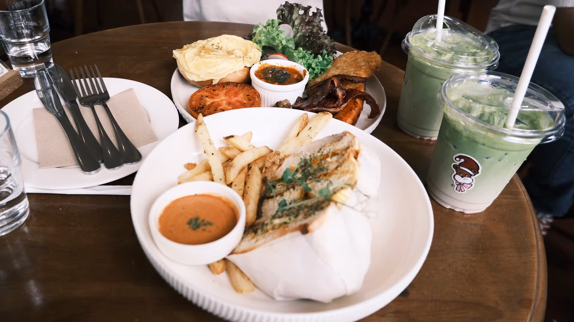 Two iced matcha drinks alongside bagel plates with fries and dipping sauce on a wooden café table at Grumpy Bagels.