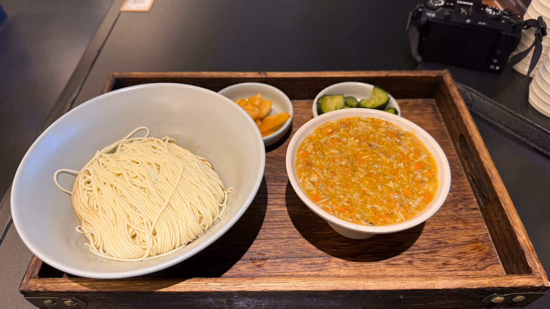 A set of hand-pulled noodles with crab broth and sides on a wooden tray, a camera visible in the background.