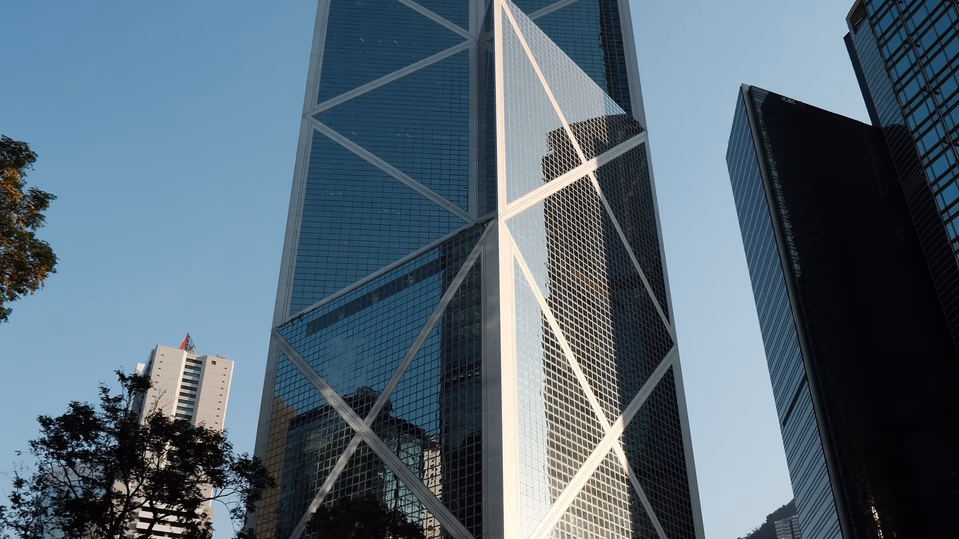 The Bank of China Tower in Central shot from street level, its geometric glass facade cutting sharply into the blue sky.