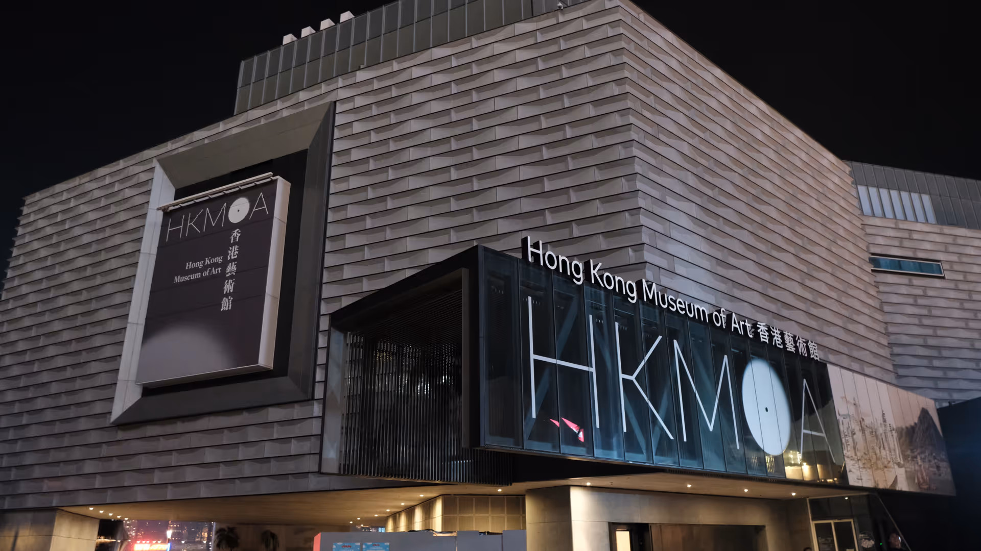 The Hong Kong Museum of Art at night, its illuminated HKMOA signage glowing on the waterfront facade.