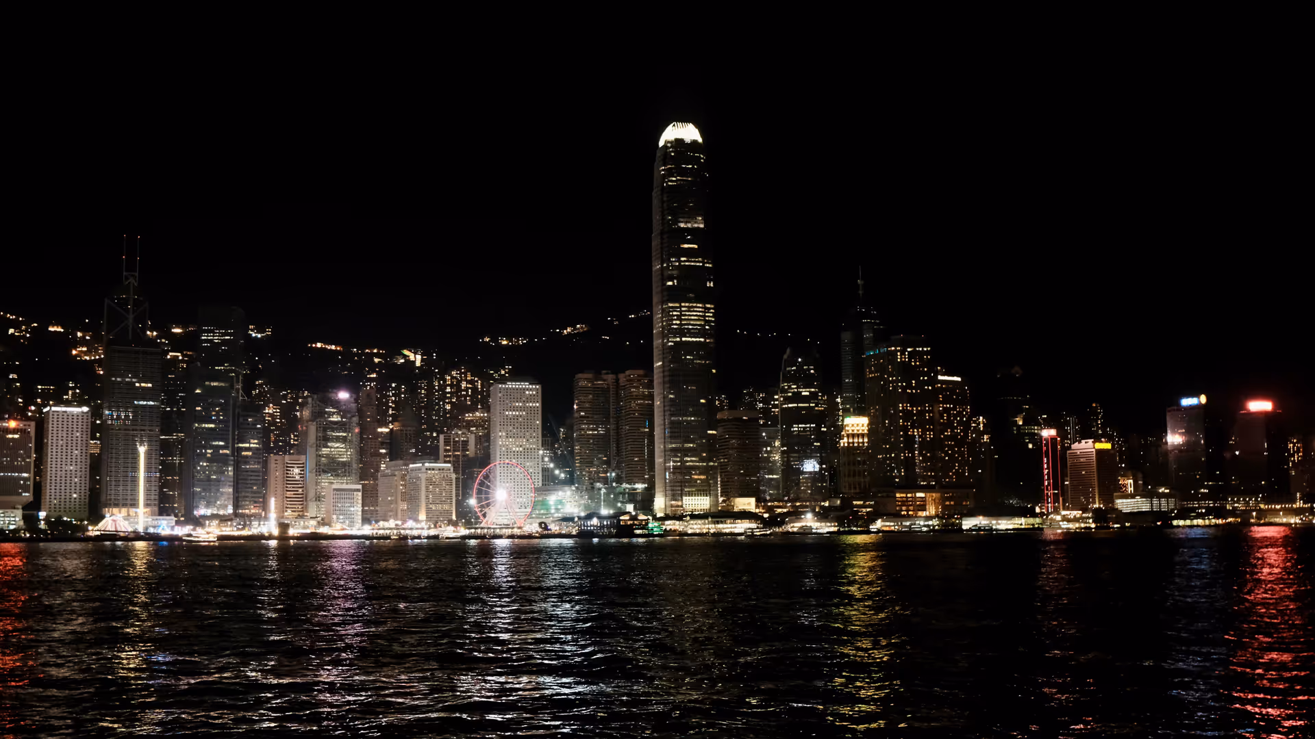 The Hong Kong Island skyline at night seen from Tsim Sha Tsui, towers reflected on the dark water of Victoria Harbour.