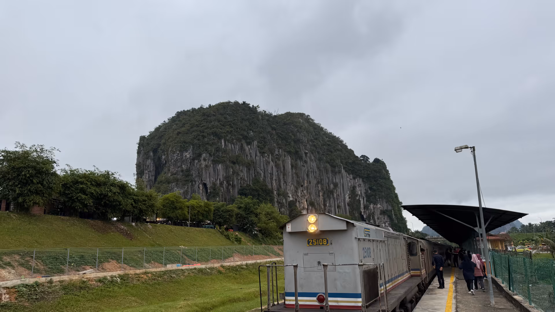 A silver KTM train engine stopped at a small rural station, framed by a massive limestone cliff.