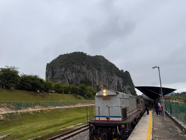 A silver KTM train engine stopped at a small rural station, framed by a massive limestone cliff.