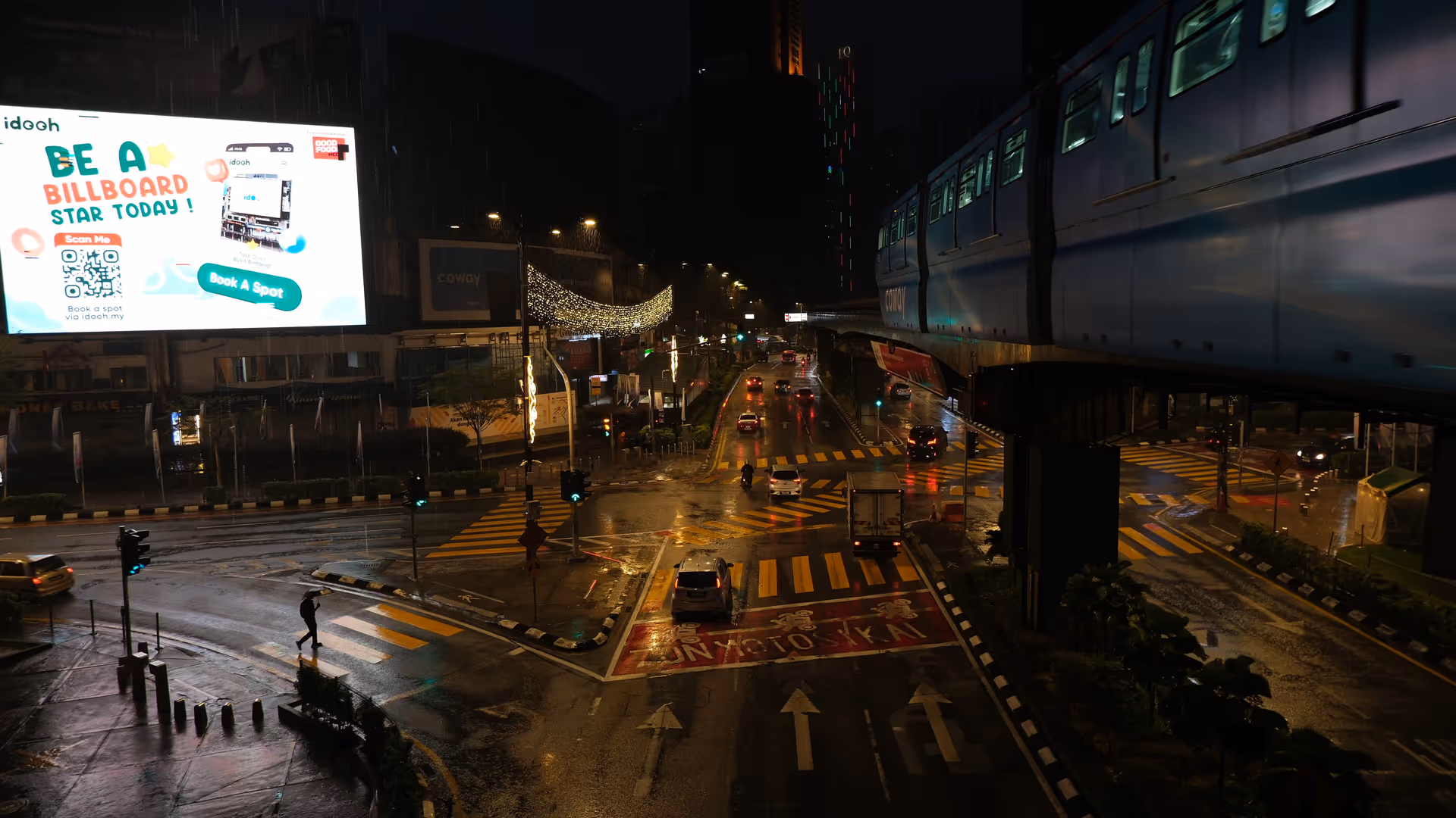 A KL monorail passing overhead at a wet nighttime intersection, with a lone pedestrian crossing below.