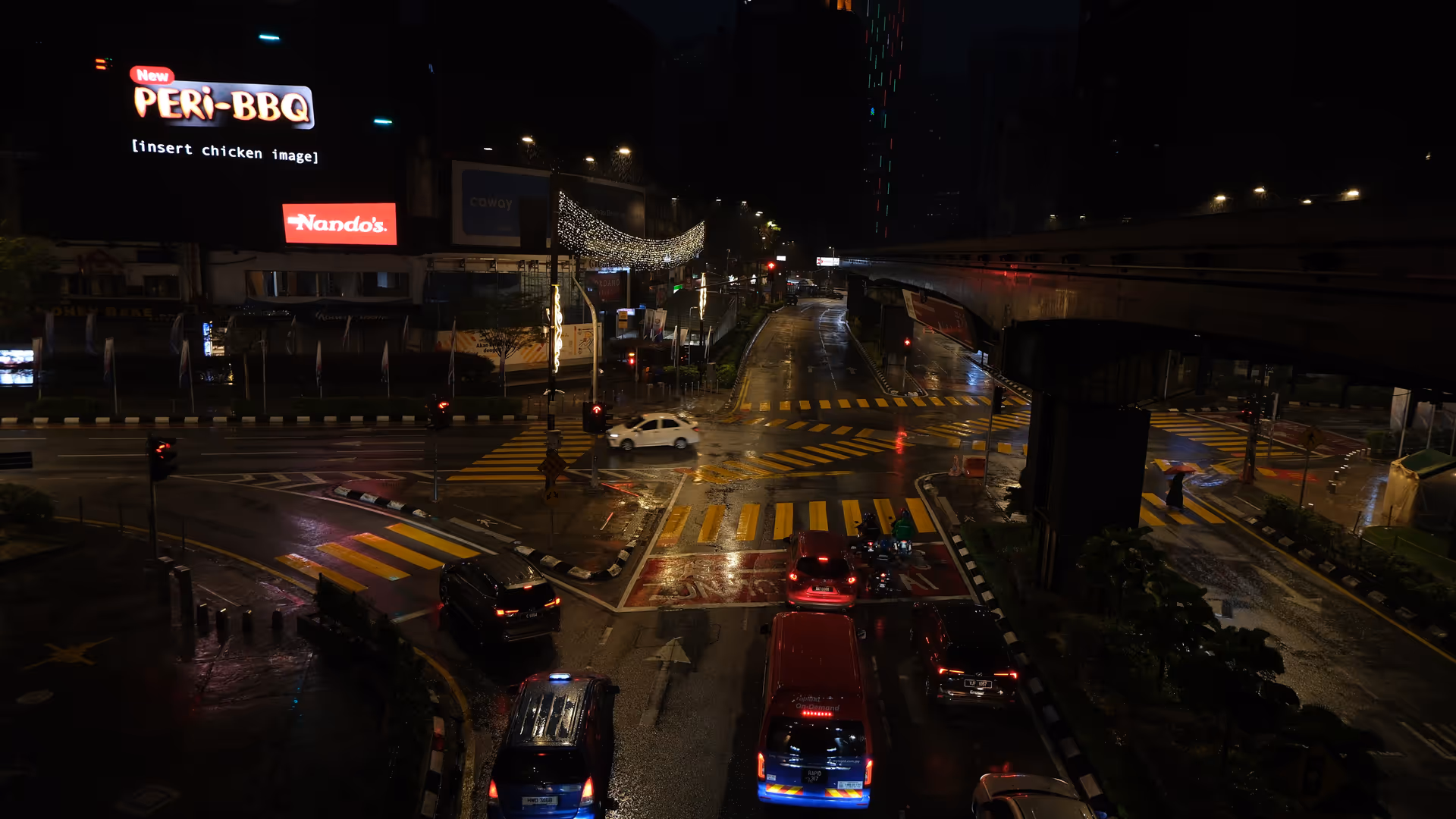 A rain-soaked KL intersection at night, with a Nando's billboard glowing above wet roads and red traffic lights.