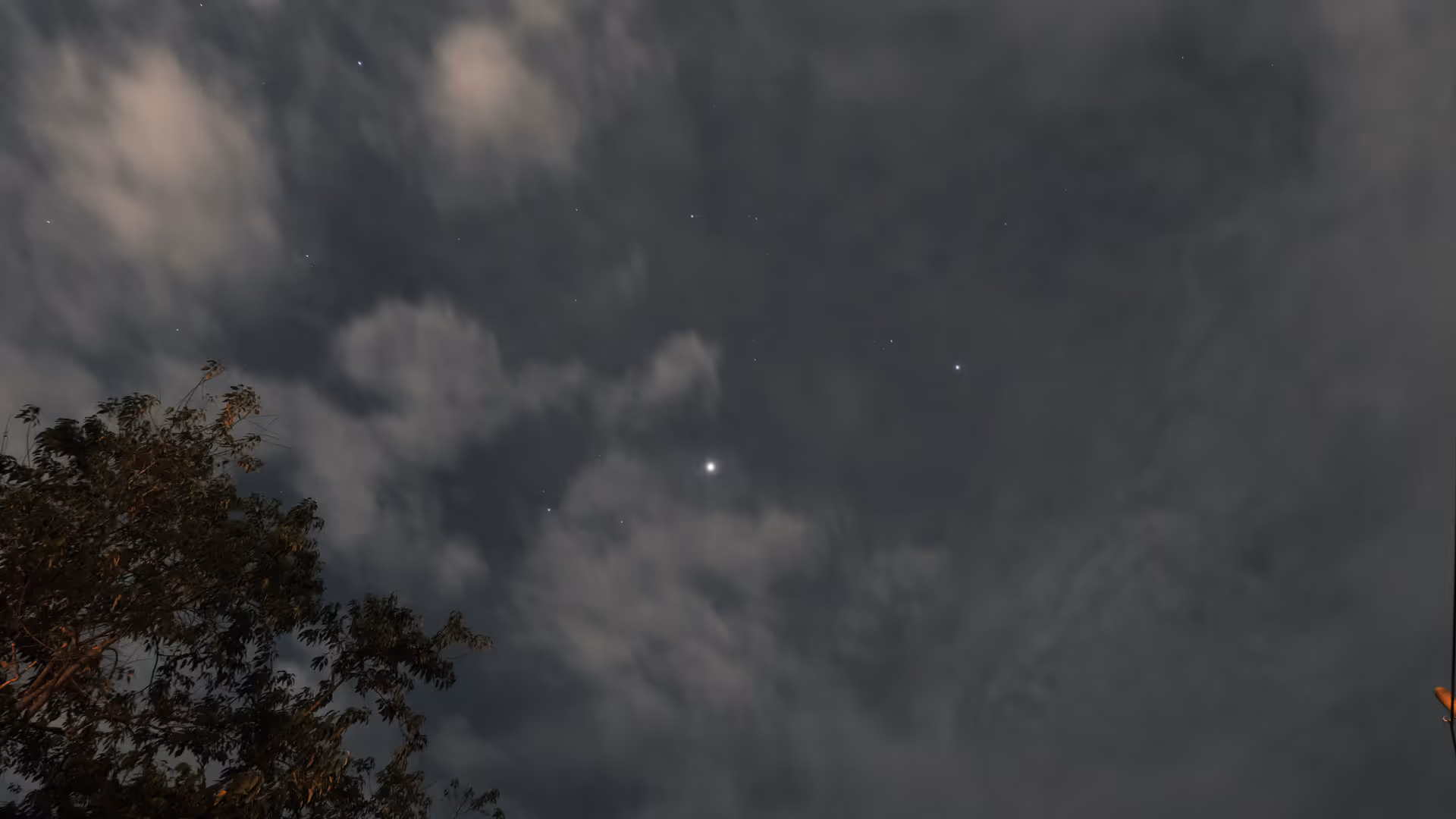 A long-exposure night sky shot showing thick cloud cover with only a handful of faint stars peeking through, a tree silhouetted in the corner.