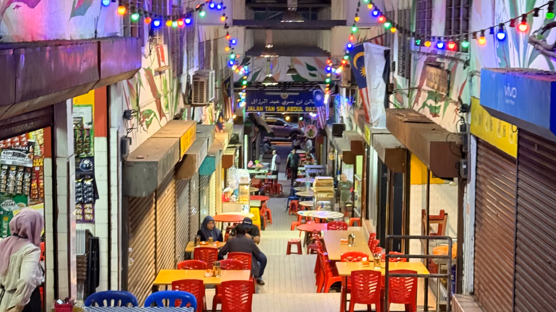 A narrow alleyway lit by colorful string lights with red and blue dining tables.
