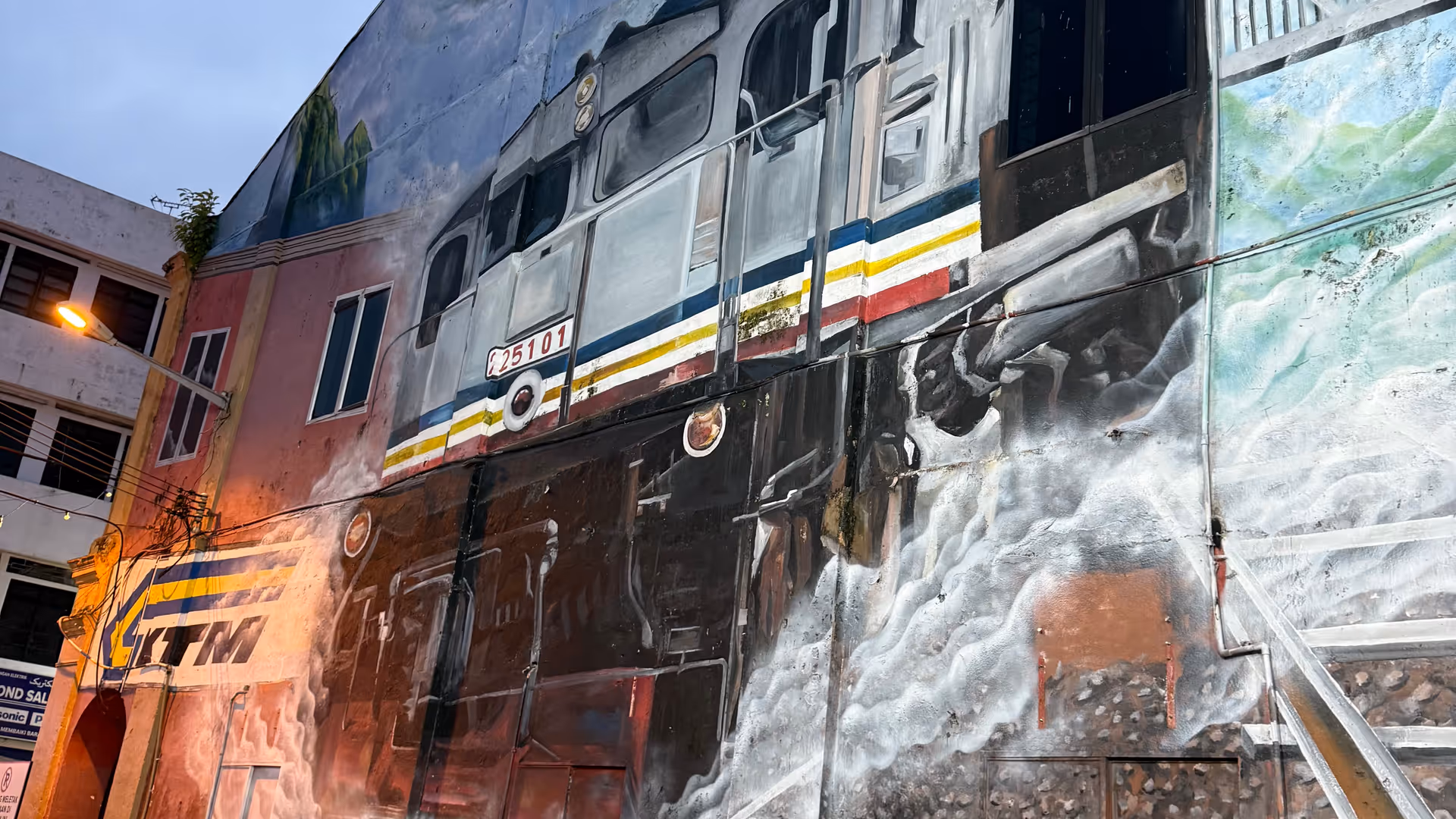 A large mural on a building side in Kuala Lipis depicting a train and a station sign.