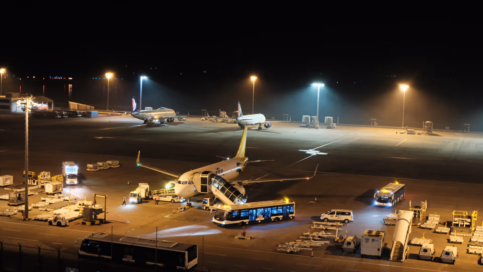 A wide-angle view of Macao Airport's tarmac at night, planes and ground crew lit up under bright floodlights.