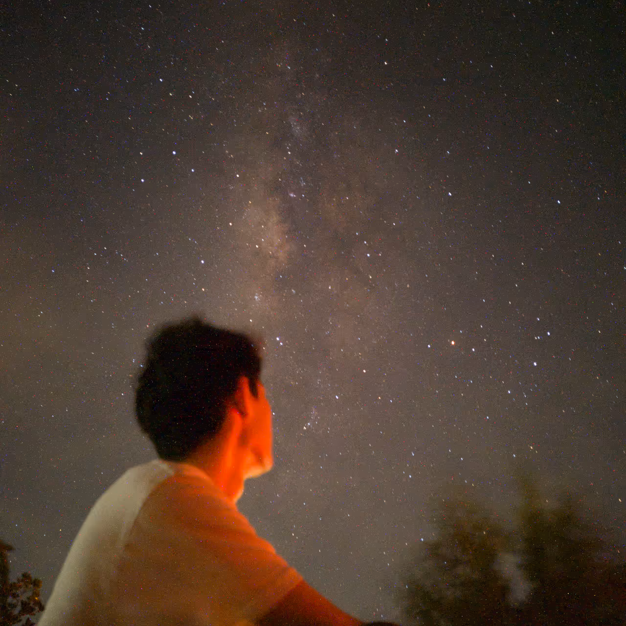 A low-angle, long-exposure silhouette of Zhun Hao sitting under a brilliant Milky Way galaxy, his face illuminated by a warm orange glow against a vast, star-filled night sky.