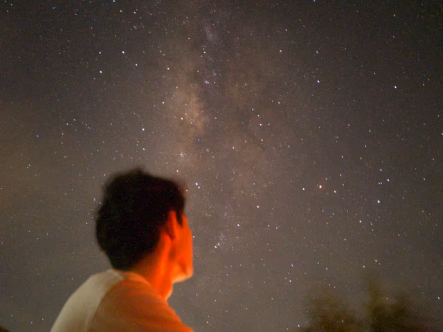 A low-angle, long-exposure silhouette of Zhun Hao sitting under a brilliant Milky Way galaxy, his face illuminated by a warm orange glow against a vast, star-filled night sky.