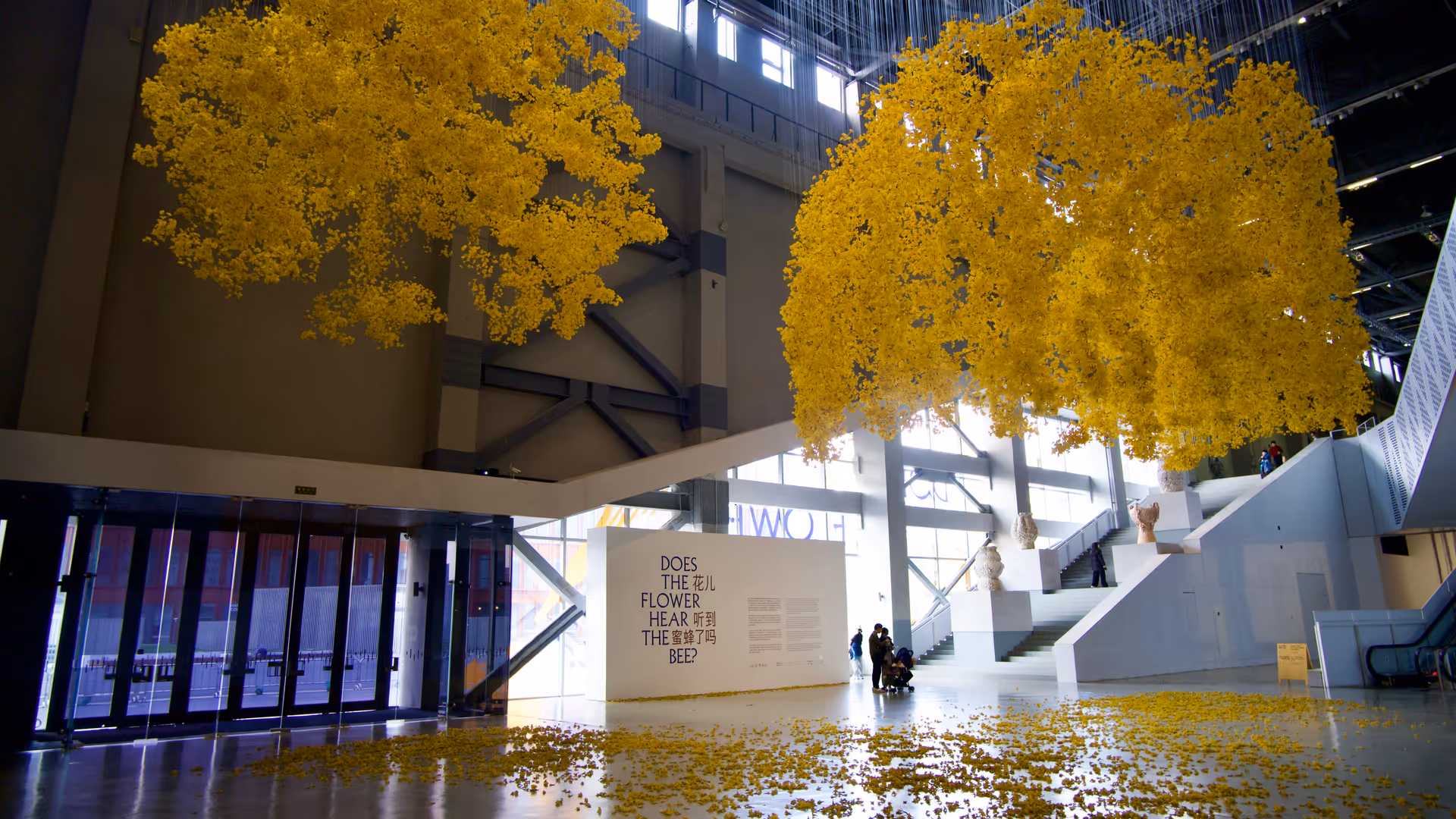 Two large upside-down yellow trees suspended from the ceiling of the Power Station of Art, their fallen leaves scattered across the gallery floor.