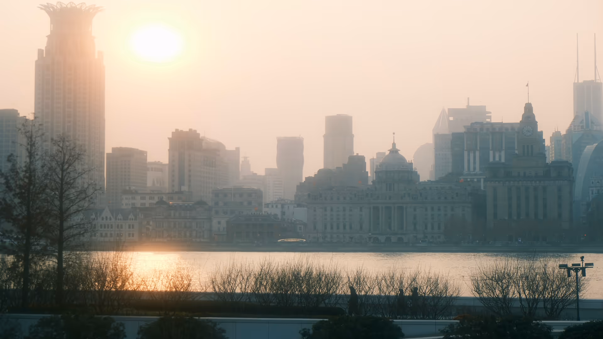 The Bund skyline seen from Pudong through a warm golden haze at sunset, the sun low behind the colonial buildings.
