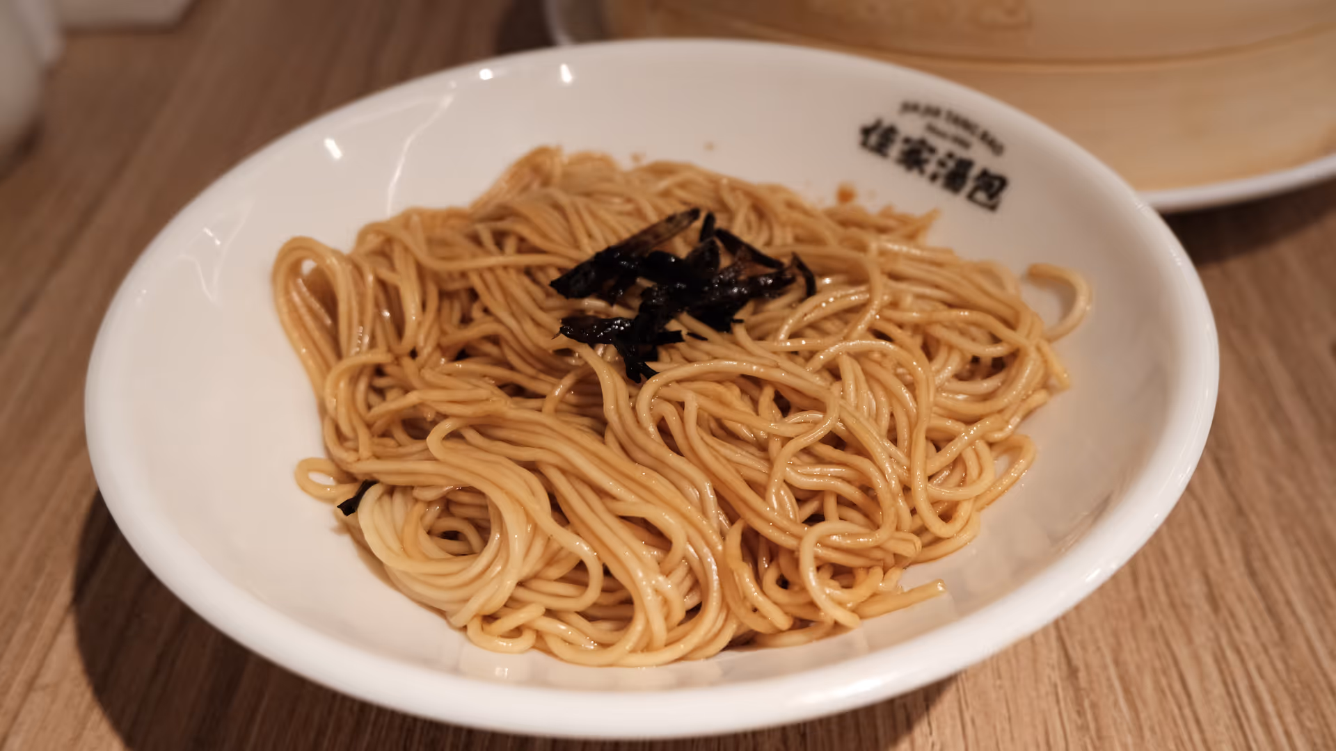 A bowl of Shanghai scallion noodles, thin and glossy, with a bamboo steamer visible in the background.
