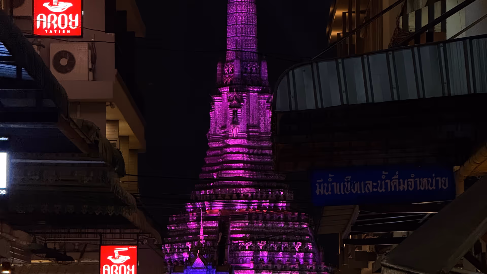 The Wat Arun spire glowing bright purple, framed by buildings on a narrow side street.