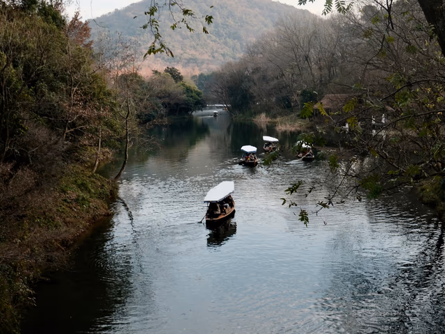 Traditional wooden boats gliding through a narrow canal at West Lake, hills and bare trees reflected in the still water.