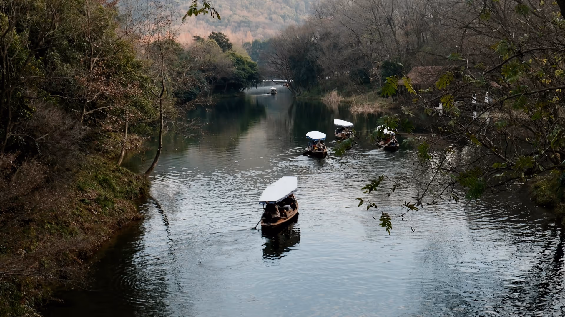 Traditional wooden boats gliding through a narrow canal at West Lake, hills and bare trees reflected in the still water.