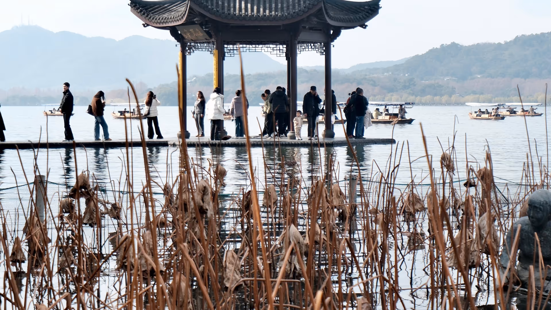 A traditional multi-tiered Chinese pavilion standing over West Lake, framed by dried winter lotus stalks in the foreground.
