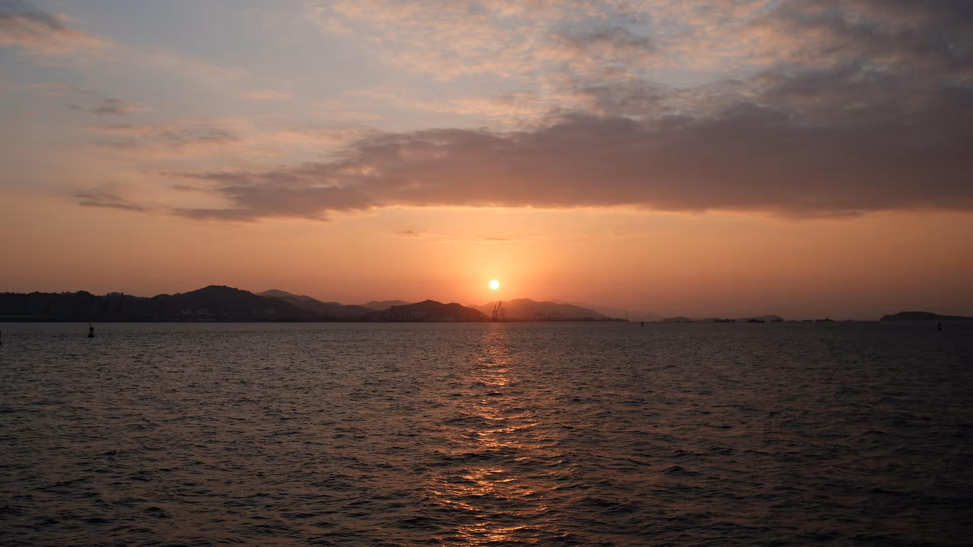 A warm orange sunset over the Xiamen Strait, the sun sinking behind distant hills reflected on calm water.
