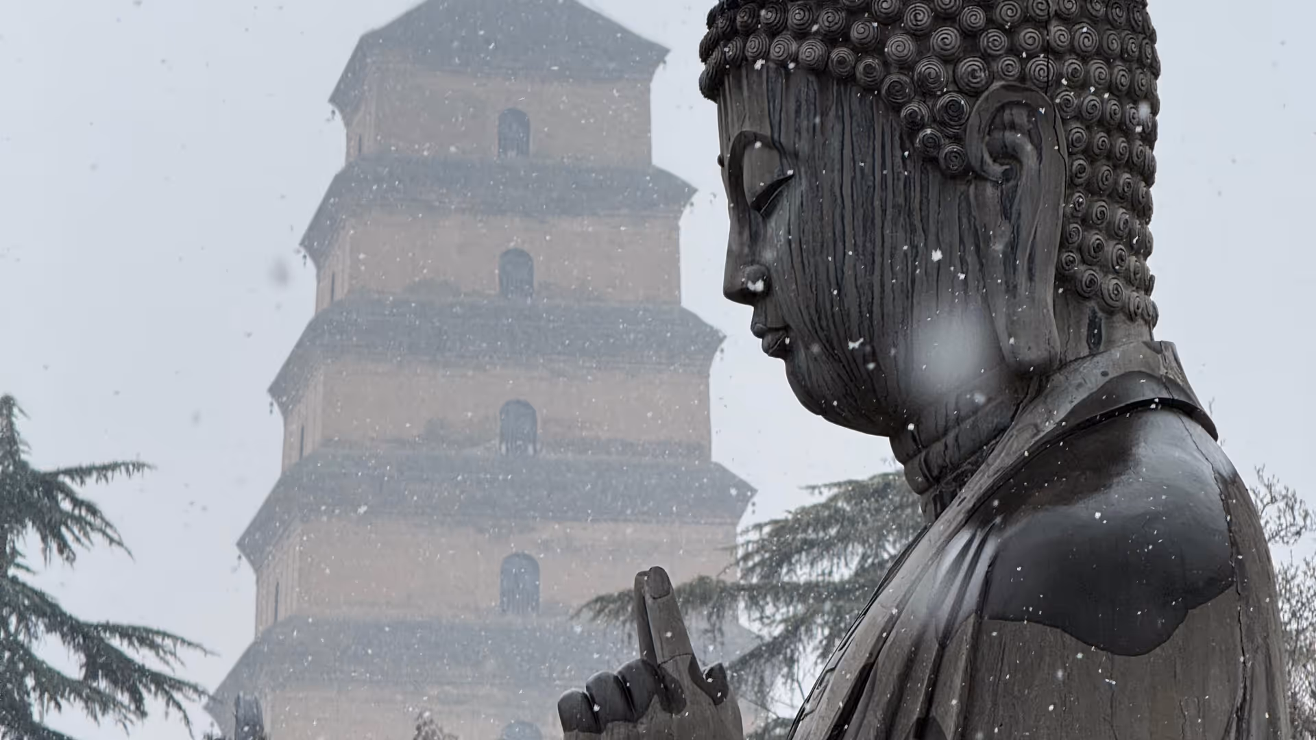 A large black Buddha statue in a blizzard, a snow-blurred pagoda visible behind it.
