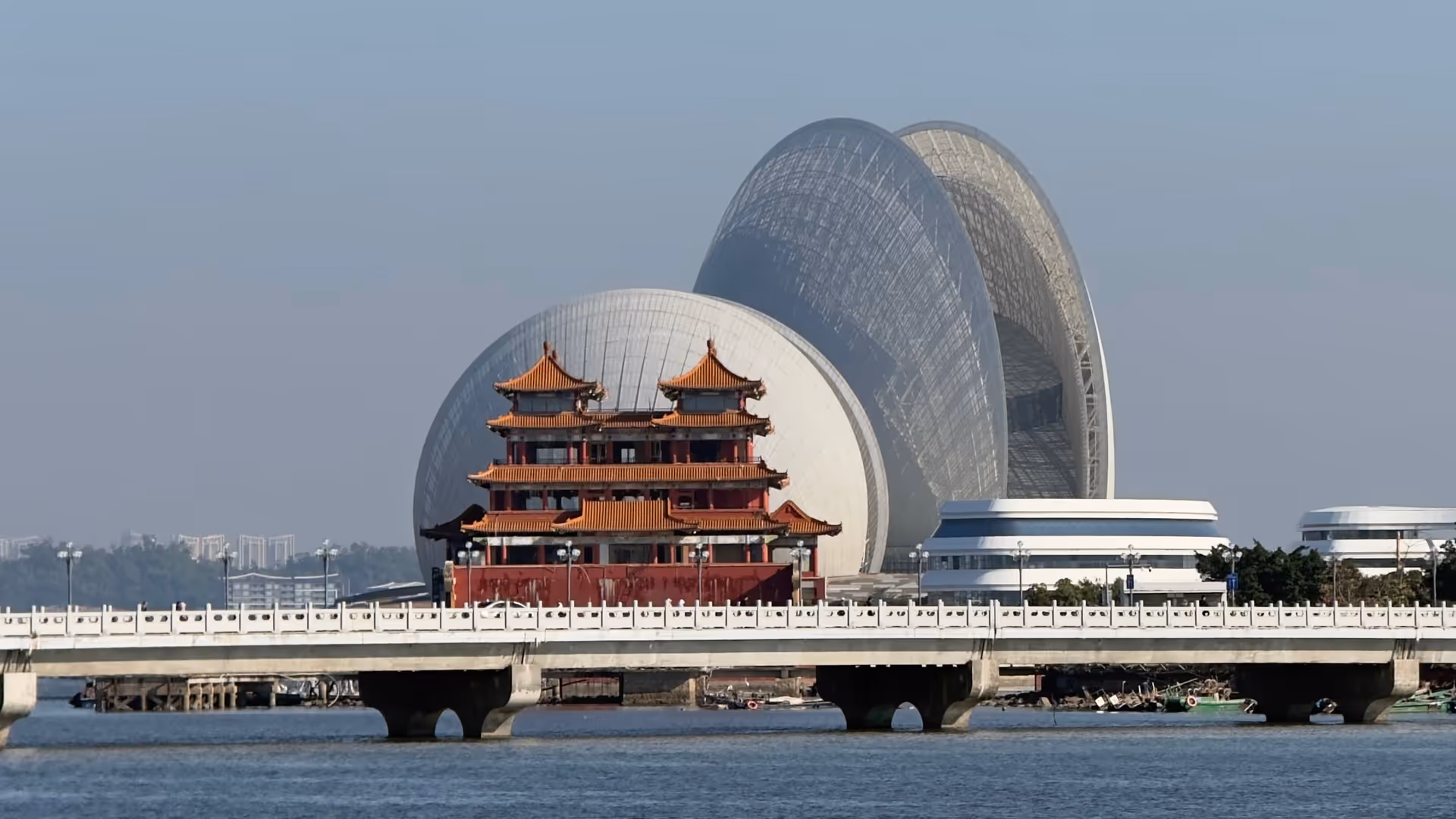 A traditional red Chinese pagoda on a bridge in the foreground, with the two sweeping oval shells of Zhuhai Grand Theatre rising behind it across the water.